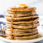 A stack of fluffy gluten-free banana oat pancakes with maple syrup and fresh banana slices on a plate.